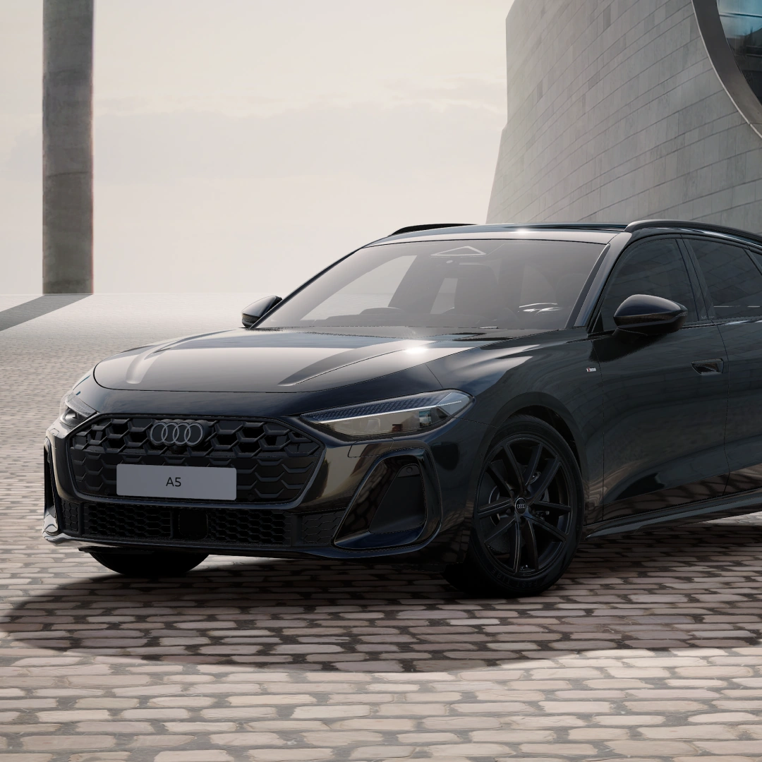 Front three-quarter view of a black Audi A5 Avant parked in a contemporary architectural setting, showcasing the Audi Singleframe grille, LED headlights, alloy wheels and sporty estate design.
